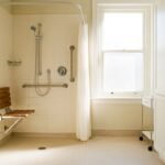 Salle de bain accessible senior, douche de plain-pied avec barre d'appui et siège, carrelage clair, lumineuse